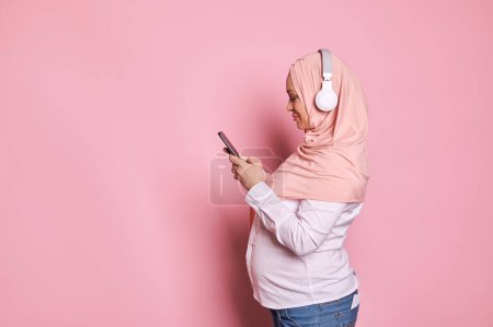 Photo for Side portrait of happy Arab Muslim woman in pink hijab with headphones and mobile phone, isolated on color background. People and technology. Mobile apps. Online communications. Pregnancy. Maternity - Royalty Free Image