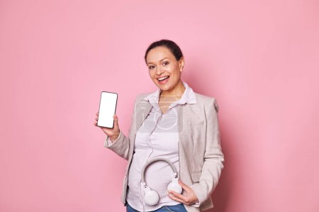 Photo for Pregnant woman holding headphones at her belly, showing white blank screen on smartphone with advertising space for insert your mobile application, smiles looking at camera, isolated pink background - Royalty Free Image
