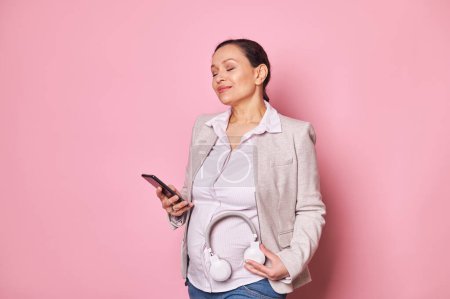 Photo for Happy pregnancy concept. Beautiful pregnant woman with her eyes closed, sharing soothing music with her upcoming new baby putting headphones on her belly, posing with smartphone on pink background - Royalty Free Image