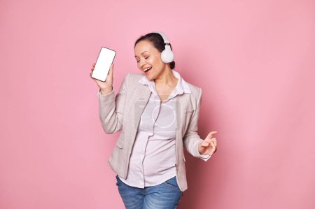 Photo for Beautiful middle-aged pregnant woman in headphones, dances to music on pink backdrop, holding a modern smartphone with white blank screen with free advertising space for insert your mobile application - Royalty Free Image