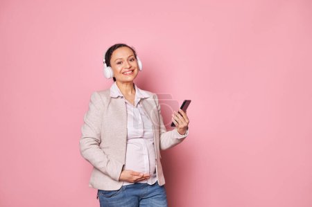 Photo for Beautiful delightful pregnant multi-ethnic woman in wireless headphones, enjoys a soothing music, smiling posing with a smartphone, scrolling playlist with cool tracks, isolated on pink background - Royalty Free Image