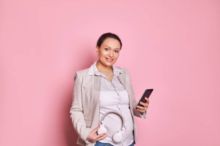 Photo for Happy pregnant woman putting headphones on her belly, puts some classical music on the playlist in mobile application on her smartphone, smiling cutely looking at camera, isolated on pink background - Royalty Free Image