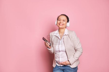 Photo for Happy pregnant multi-ethnic woman in wireless headphones, checking new mobile application on smartphone, rejoicing at feeling first baby kicks, smiling looking at camera over pink background - Royalty Free Image