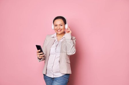 Photo for Young multi-ethnic pregnant woman in casual clothes, smiling with beautiful toothy smile looking at camera listening to music in wireless headphones and holding smartphone on isolated pink background - Royalty Free Image