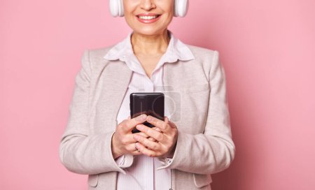 Photo for Details on the hands of a blurred smiling woman using smartphone, scrolling playlist, checking mobile apps, enjoying listening to music in headphones, isolated over pink background. Copy space - Royalty Free Image