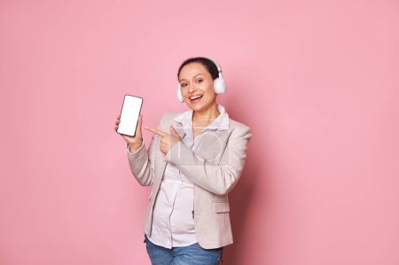Photo for Attractive pregnant woman in headphones, smiling looking at camera, showing smartphone with blank white screen with copy space for advertising text or mobile application, isolated on pink background - Royalty Free Image