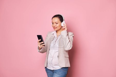 Photo for Attractive multi-ethnic middle-aged pregnant woman wearing wireless headphones, listening to music or watching newborn baby care video on the internet, isolated pink background. Maternity. Pregnancy - Royalty Free Image