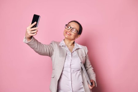Photo for Attractive middle-aged multi-ethnic pregnant woman, business person wearing casual clothes and trendy glasses, carrying laptop and taking selfie on her smartphone on isolated pink background. Ad space - Royalty Free Image