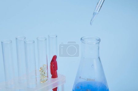 Téléchargez les photos : Concentration sélective sur les gouttes avec une solution chimique bleue coulant d'une pipette graduée dans une fiole de laboratoire, près des éprouvettes avec des capsules de gel sur fond bleu isolé. Chimie et pharmacie - en image libre de droit