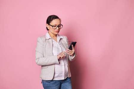 Photo for Confident middle-aged ethnic pregnant business woman, dressed in casual wear, checking a pregnancy mobile application on smartphone, counting baby kicks, browsing websites, isolated on pink background - Royalty Free Image
