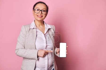 Photo for Happy pregnant woman showing you a smartphone with blank white digital screen with mockup for mobile application, smiling looking at camera, isolated pink background. Copy space for advertising text - Royalty Free Image