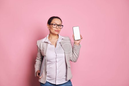 Photo for Multi ethnic confident pretty pregnant business woman wearing trendy glasses, carrying laptop, showing a smartphone with white blank digital screen, pink background. Mockup. Copy space for mobile apps - Royalty Free Image