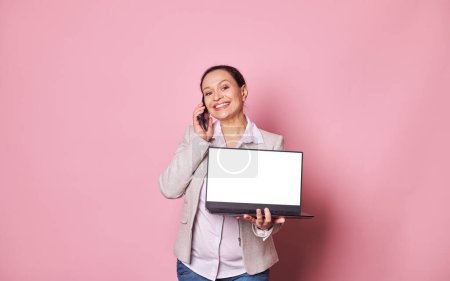 Photo for Pleasant pregnant woman, sales manager, entrepreneur talking on mobile phone and showing at camera a laptop with white screen with copy space for mobile apps, smiling isolated over pink background - Royalty Free Image