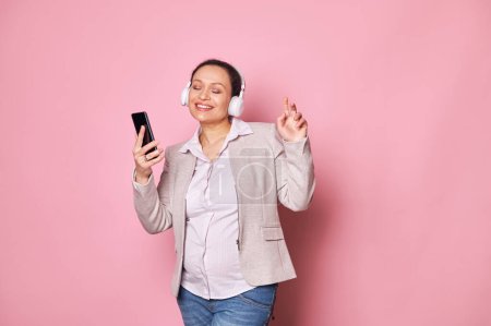Photo for Happy delightful pregnant woman enjoying soothing music in wireless headphones, holding mobile phone, smiling and dancing, isolated on pink background. Carefree pregnancy concept. Copy ad space - Royalty Free Image