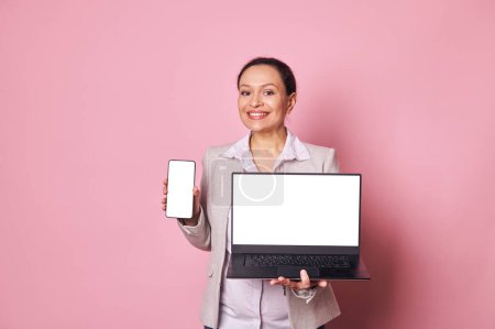 Photo for Pleasant pregnant business woman showing you a modern smartphone and laptop, with white blank digital screen with copy space for your ads or mobile applications, smiling over pink background. Mockup - Royalty Free Image