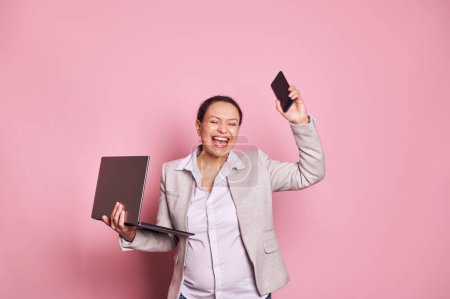 Photo for Overjoyed pregnant middle-aged business woman, dressed in casual clothing, smiling and expressing excitement and joy, posing with her eyes closed over pink background, holding laptop and mobile phone - Royalty Free Image