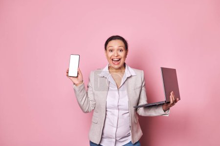 Photo for Excited pregnant business lady expressing amazement, looking at camera, holding laptop and smartphone with white blank screen, with free ad space for mobile applications, isolated over pink background - Royalty Free Image