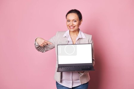 Photo for Adorable multi ethnic working pregnant woman smiles looking at camera, points her finger at white blank digital screen on laptop, isolated pink background. Copy ad space. Mockup. Pregnancy and career - Royalty Free Image