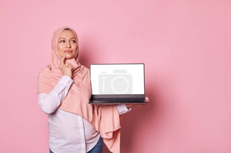 Photo for Pensive pregnant Muslim woman in pink hijab, holds laptop with white blank screen, looking thoughtfully aside, reasoning over business project, planning her maternity leave, isolated pink background - Royalty Free Image