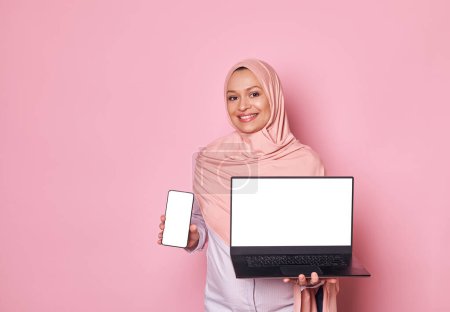 Photo for Arab Muslim pretty woman in pink hijab, smiling looking at camera, showing digital gadgets - a laptop and smartphone with white blank screen. Copy space for mobile apps, isolated on pink background - Royalty Free Image