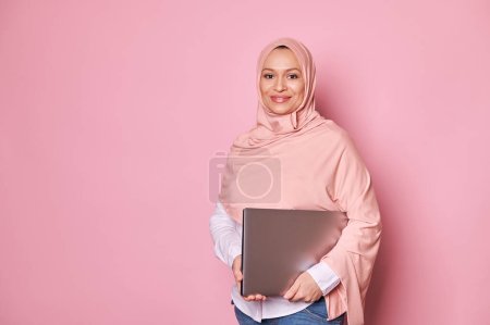 Confident successful beautiful Arab Muslim female manager, business lady, entrepreneur, working pregnant woman in pink hijab, holding laptop, smiling cutely looking at camera, isolated pink background
