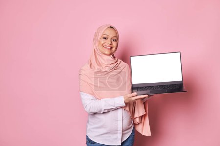 Photo for Confident pregnant Arab Muslim woman with head covered in pink hijab, smiling looking at camera, holding laptop with white blank digital screen. Mockup for insert mobile app, isolated pink background - Royalty Free Image