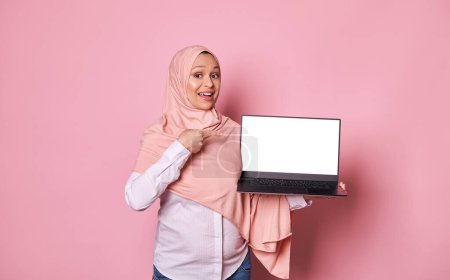 Photo for Middle-Eastern Muslim woman, working pregnant female entrepreneur, sales manager dressed in pink hijab, points her finger at copy space for ads on laptops white empty screen, isolated pink background - Royalty Free Image
