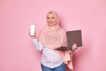 Photo for Smiling charming pregnant Arab Muslim woman in pink hijab, looking at camera, showing a smartphone with white blank screen with free ad space, isolated pink background. Mobile apps. People. Technology - Royalty Free Image
