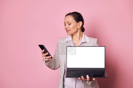 Photo for Attractive confident pregnant business woman checking mobile application on her smart phone, holding laptop with white blank screen with free advertising space, isolated on pink color background - Royalty Free Image