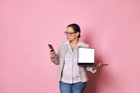 Photo for Multitasking pregnant woman checking smartphone over pink background, holding a laptop with white empty screen, with copy space for ads or mobile applications. People. Pregnancy. Business. Technology - Royalty Free Image