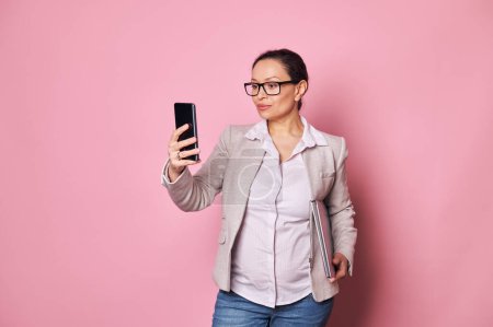 Photo for Pregnant confident Hispanic business woman wearing casual clothes and stylish glasses, holding laptop, checking mobile app on her smartphone, isolated on pink background. Career and maternity concept - Royalty Free Image