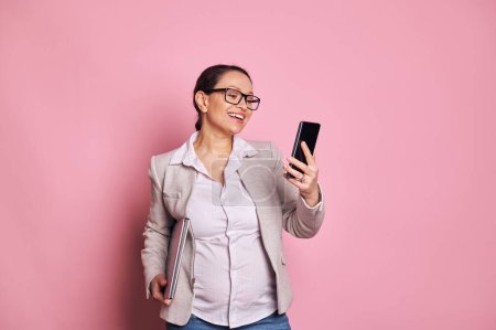 Photo for Portrait of middle aged confident multi ethnic happy pregnant business woman, wearing trendy glasses and casual wear, holding laptop, using mobile phone on isolated pink background. People and career - Royalty Free Image