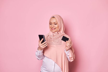 Photo for Charming Arab Muslim pregnant woman in pink hijab, holding credit card and smartphone, makes cashless payments via internet mobile banking, isolated background. Online shopping Booking Ordering food - Royalty Free Image