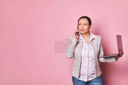 Photo for Multitasking business lady, working pregnant woman talking on mobile phone, negotiating with investors, holding laptop, isolated over pink background with copy ad space. Pregnancy and career concept - Royalty Free Image