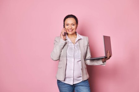 Photo for Stylish pregnant multi ethnic woman, businesswoman talking on mobile phone, negotiating with business partners, concluding deals, smiling looking at camera, posing with laptop, isolated pink backdrop - Royalty Free Image