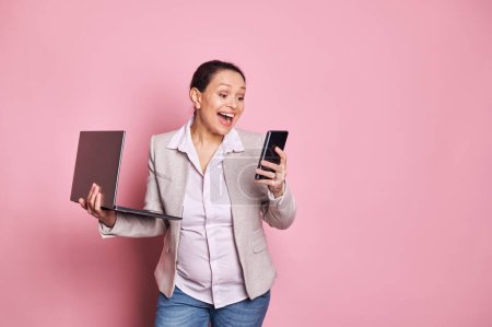 Photo for Excited happy working pregnant multi ethnic woman checking her smartphone and laptop, expressing joy, looking forward to maternity leave, on pink isolated background. Pregnancy and career concept - Royalty Free Image
