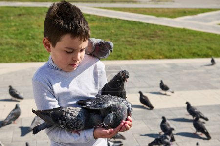 Téléchargez les photos : Adorable garçon multi ethnique de 10 ans, nourrissant des pigeons sur la place de la ville par une journée ensoleillée. Amour et soin des animaux. Concept des personnes et de la nature - en image libre de droit