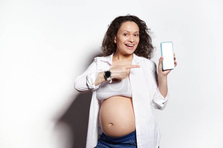 Photo for Smiling curly haired multi ethnic pregnant woman showing modern smartphone with white blank digital screen with free space for ads or mobile apps on isolated background. Pregnancy 2nd semester. Mockup - Royalty Free Image