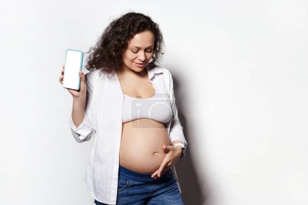 Photo for Delightful pregnant woman in 30 week of pregnancy, gently caressing her belly, holding mobile phone with white blank digital display, isolated background. Maternity. People and technology concept - Royalty Free Image