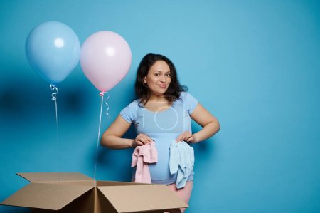 Happy pregnant woman with big belly, holding newborn clothes, smiling at camera, posing with balloons of pink and blue pastel color, isolated blue background. Baby shower. Gender reveal party concept