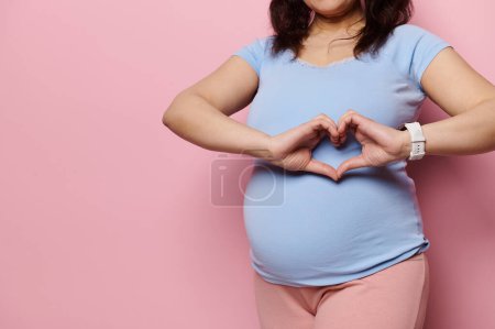 Focus on the hands of a pregnant woman, expectant gravid mother in blue t-shirt, showing heart shape made with her fingers over her big belly, expressing love, tenderness, isolated on pink background