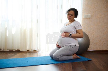 Foto de Joven mujer embarazada multi étnica hermosa, futura madre grávida practicando yoga en un ambiente hogareño tranquilo, esperando el nacimiento de su bebé. El concepto de salud y bienestar en el embarazo - Imagen libre de derechos