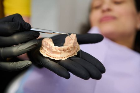 Téléchargez les photos : Dentiste montrant un moule de plâtre dentaire à la caméra, tenant la main au-dessus d'une patiente allongée sur une chaise de dentiste, recevant un traitement en clinique dentaire. Modèle de mâchoire. Concept de soins de santé, dentaires et buccodentaires - en image libre de droit
