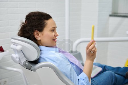 Téléchargez les photos : Portrait latéral d'une patiente heureuse dans une chaise de dentiste, souriante, regardant dans un miroir, satisfaite du traitement et du blanchiment des dents professionnel dans une clinique dentaire. Soins buccodentaires et hygiène dentaire concept - en image libre de droit