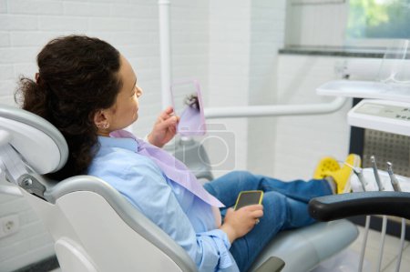 Téléchargez les photos : Femme enceinte souriante, patiente assise dans une chaise de dentiste, regardant dans le miroir après un traitement dentaire professionnel dans une clinique dentaire. Pratique dentaire. Prévention des caries et traitement des dents - en image libre de droit