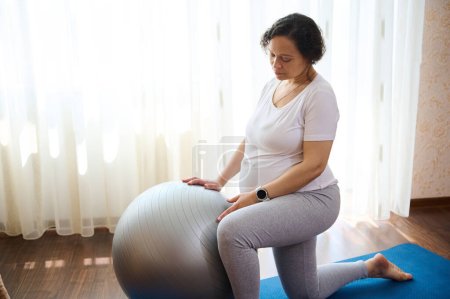 Multi ethnic adult pregnant woman exercising with a fit ball at home. Gravid expectant mother in sportswear, doing prenatal fitness exercises for wellness and wellbeing in pregnancy time and maternity