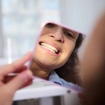 Close-up reflection in the cosmetic mirror of a pretty woman, female patient sitting in dental chair, admiring her beautiful smile and teeth after teeth bleaching procedure in dentistry clinic.