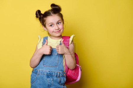 Téléchargez les photos : Adorable petite fille adorable en t-shirt jaune décontracté et salopette en denim bleu, les pouces levés, souriant, isolé sur fond de studio jaune. Copier l'espace publicitaire. L'éducation. Retour à l'école - en image libre de droit
