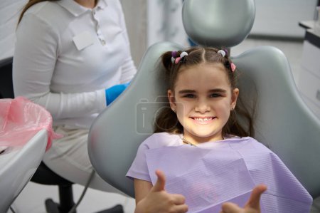 Lächelndes kleines Mädchen beim Zahnarzttermin, auf dem Zahnarztstuhl sitzend, Daumen hoch nach der Vorsorgeuntersuchung in der modernen Kinderzahnklinik. Kinderzahnheilkunde. Vorbeugung gegen Karies