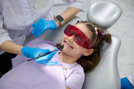 Téléchargez les photos : Jolie petite fille dans la chaise des dentistes, portant des lunettes de protection UV, souriant à un médecin dentiste tenant miroir dentaire tandis qu'un examen des dents dans la clinique de dentisterie pédiatrique. Concept de contrôle dentaire - en image libre de droit